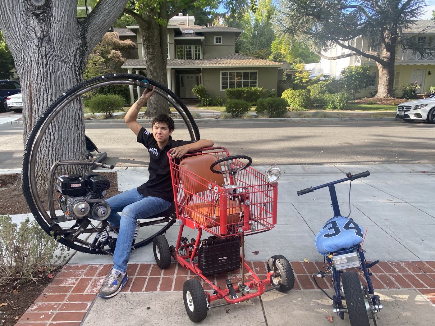 Spencer sitting on the monowheel beside the go-kart and mini-bike on a sidewalk